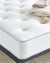 Close-up of a plush white mattress with a pillowy top layer on a dark platform bed frame, accompanied by a neatly folded beige blanket and striped pink and white pillows at the foot of the bed.