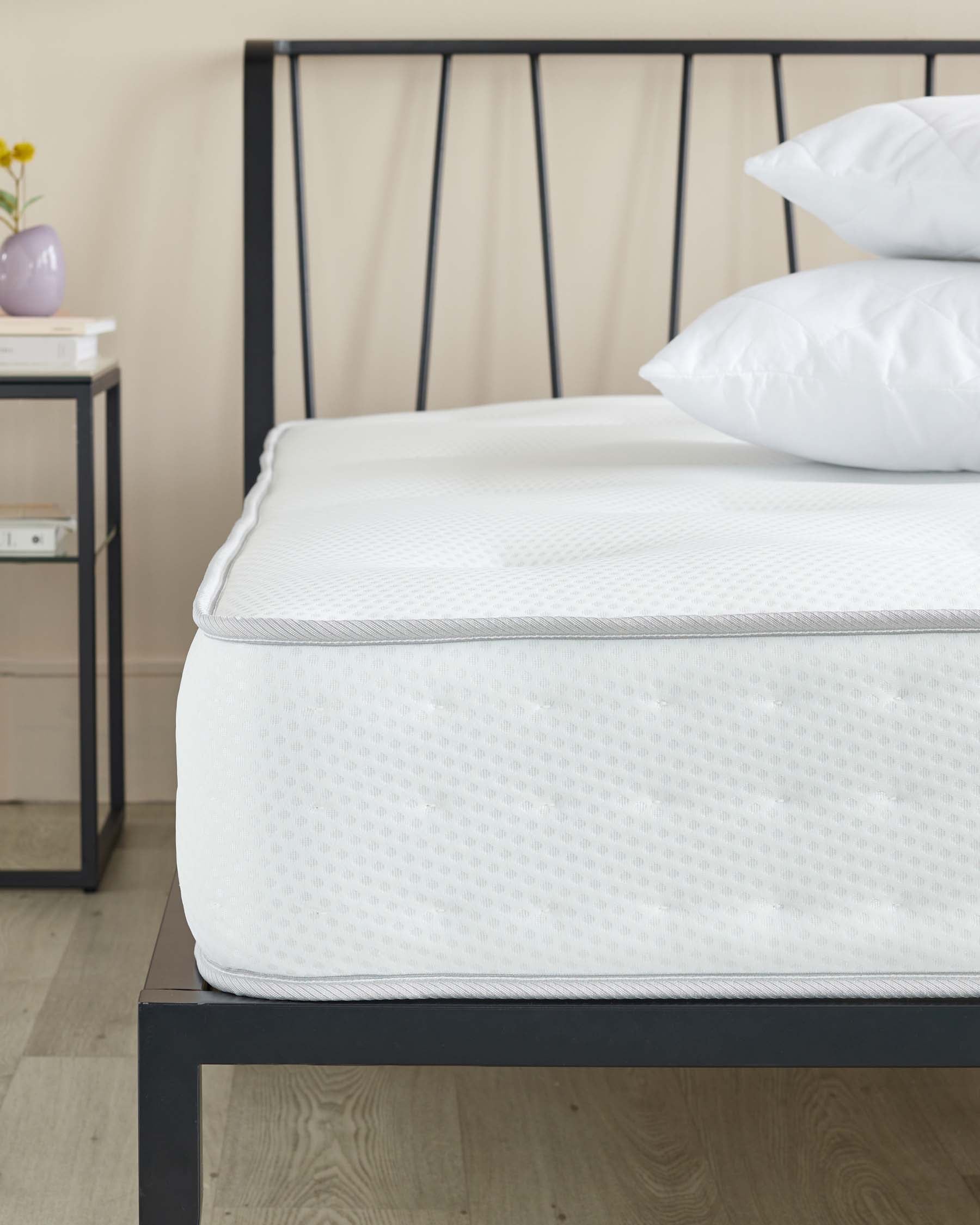 A contemporary metal bed frame with a matte black finish and an elegantly curved headboard featuring vertical spindles. The bed is partially dressed with a plush white mattress and two fluffy pillows without linens. Beside the bed is a minimalist nightstand with a pale finish and simple lines, adorned with a small vase and books.