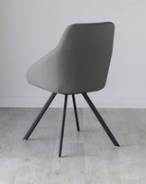 Modern grey upholstered dining chair with a curved backrest and four black metal angled legs on a light wooden floor against a white wall.