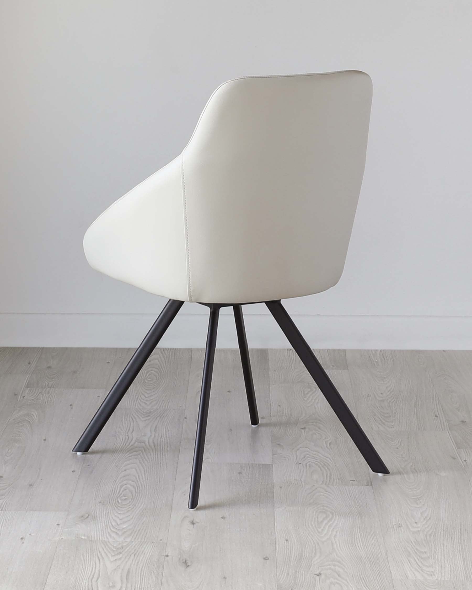 Modern minimalist style chair with a smooth cream-colored faux leather upholstery, featuring a curved backrest and a spacious seat. The chair is supported by four slanted, matte black metal legs that taper down to fine points, creating a sleek and elegant silhouette. The chair is set against a simple white wall and a light wood laminate floor, emphasizing its contemporary design.