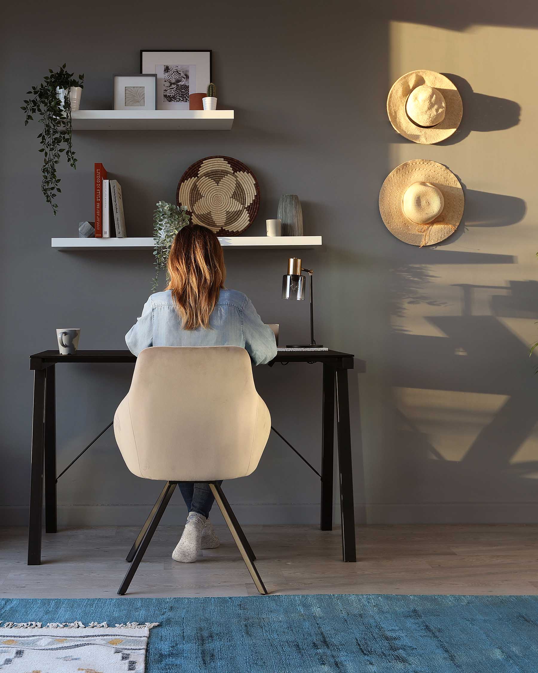 A contemporary home office setup featuring a sleek, black wooden desk with angular legs, paired with a modern, beige upholstered chair with a unique backrest design and metal legs. Above the desk, two floating white shelves display various decorative items, enhancing the clean and minimalist aesthetic.