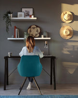 Contemporary furniture set against a grey wall, featuring a sleek black writing desk with straight lines and a simple, sturdy design. Accompanied by an ergonomic teal office chair with a curved backrest and black metal legs. Two white floating shelves above the desk display decorative items, casting soft shadows in the ambient light.