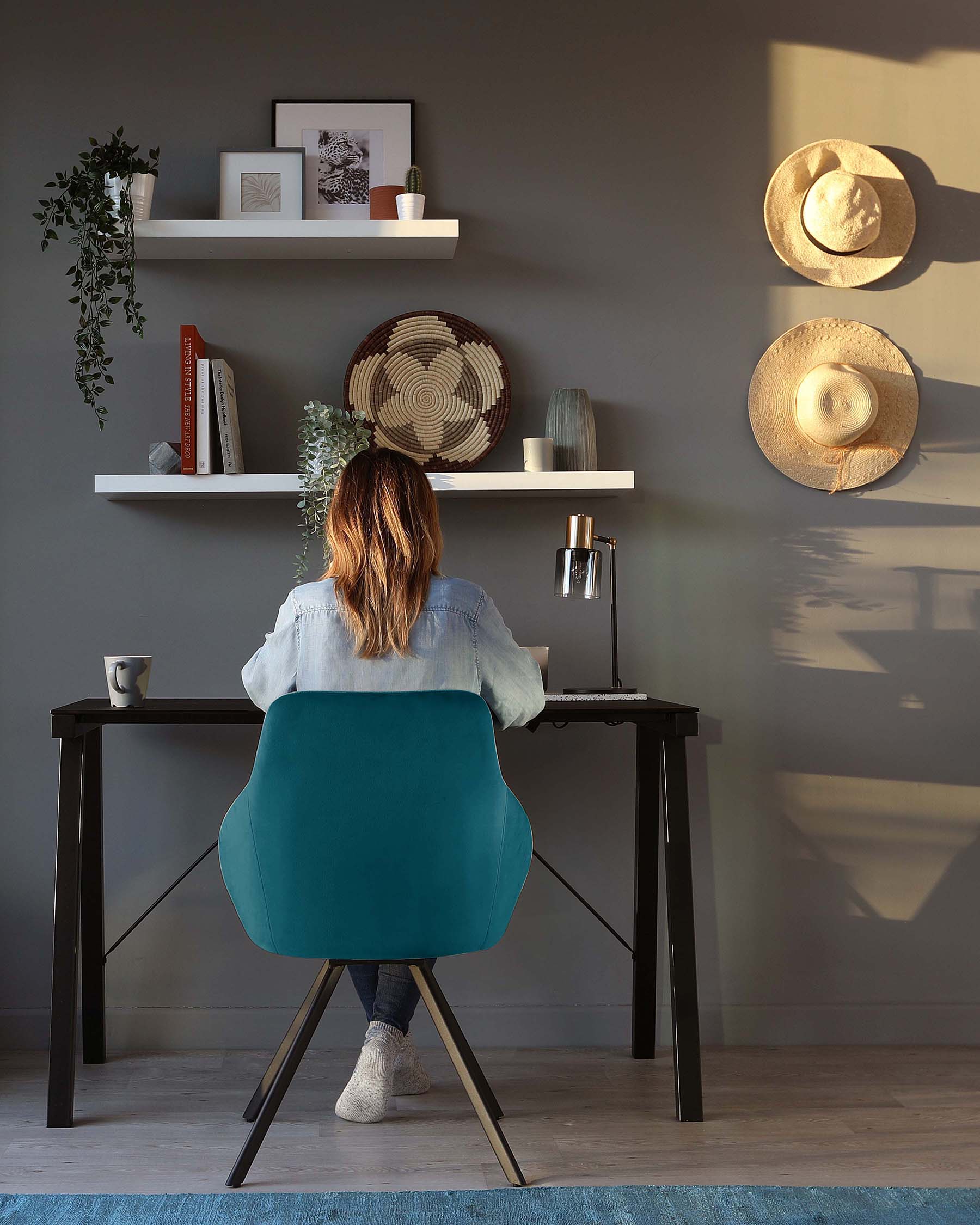 Contemporary furniture set against a grey wall, featuring a sleek black writing desk with straight lines and a simple, sturdy design. Accompanied by an ergonomic teal office chair with a curved backrest and black metal legs. Two white floating shelves above the desk display decorative items, casting soft shadows in the ambient light.