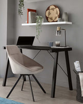 Modern home office setup featuring an A-frame black wooden desk paired with a light taupe, cushioned desk chair with black metal legs. A white floating shelf is mounted above the desk, holding decorative items.