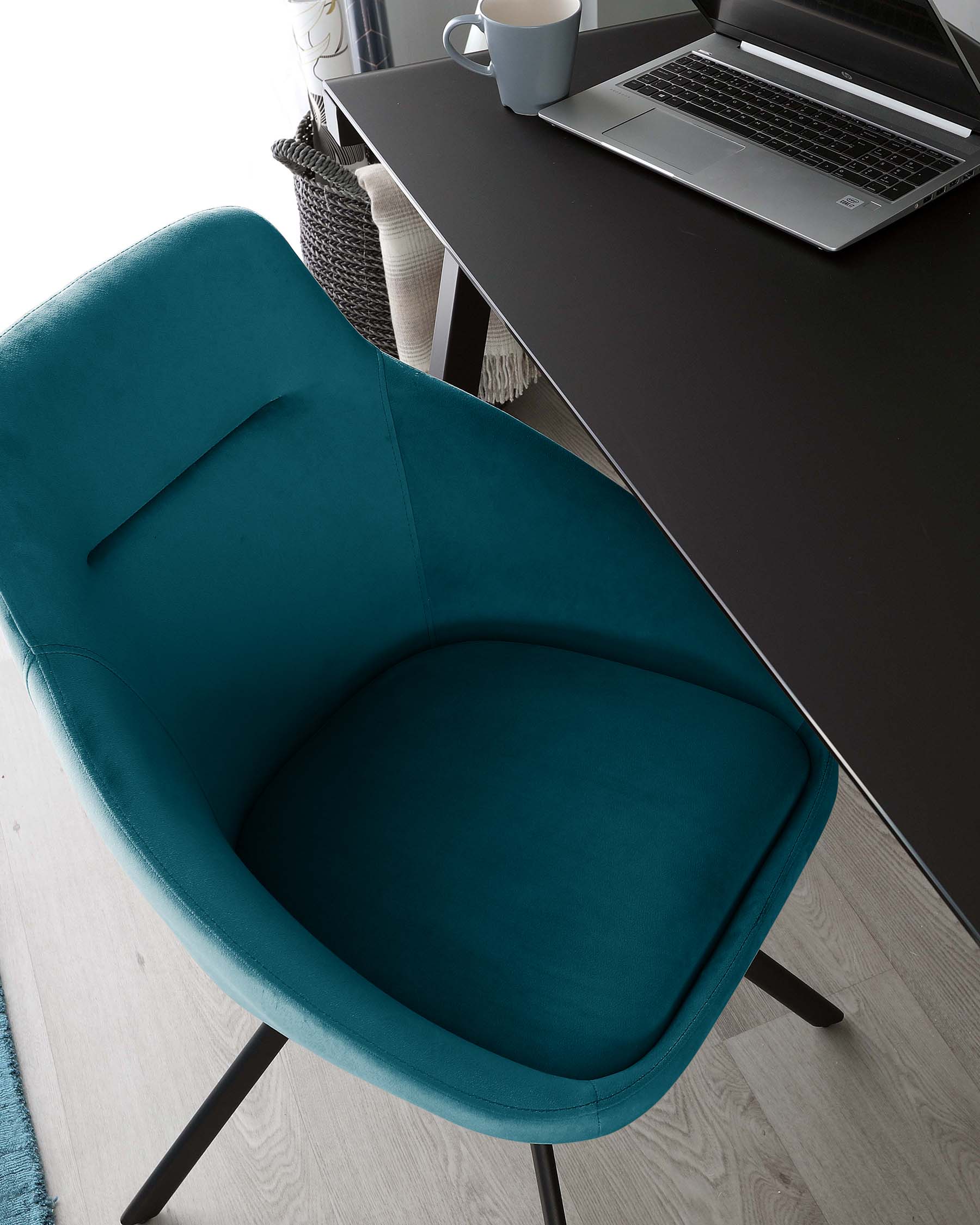 Modern teal upholstered chair with a curved backrest and sleek black metal legs, situated beside a contemporary black desk with a laptop and white mug on top.