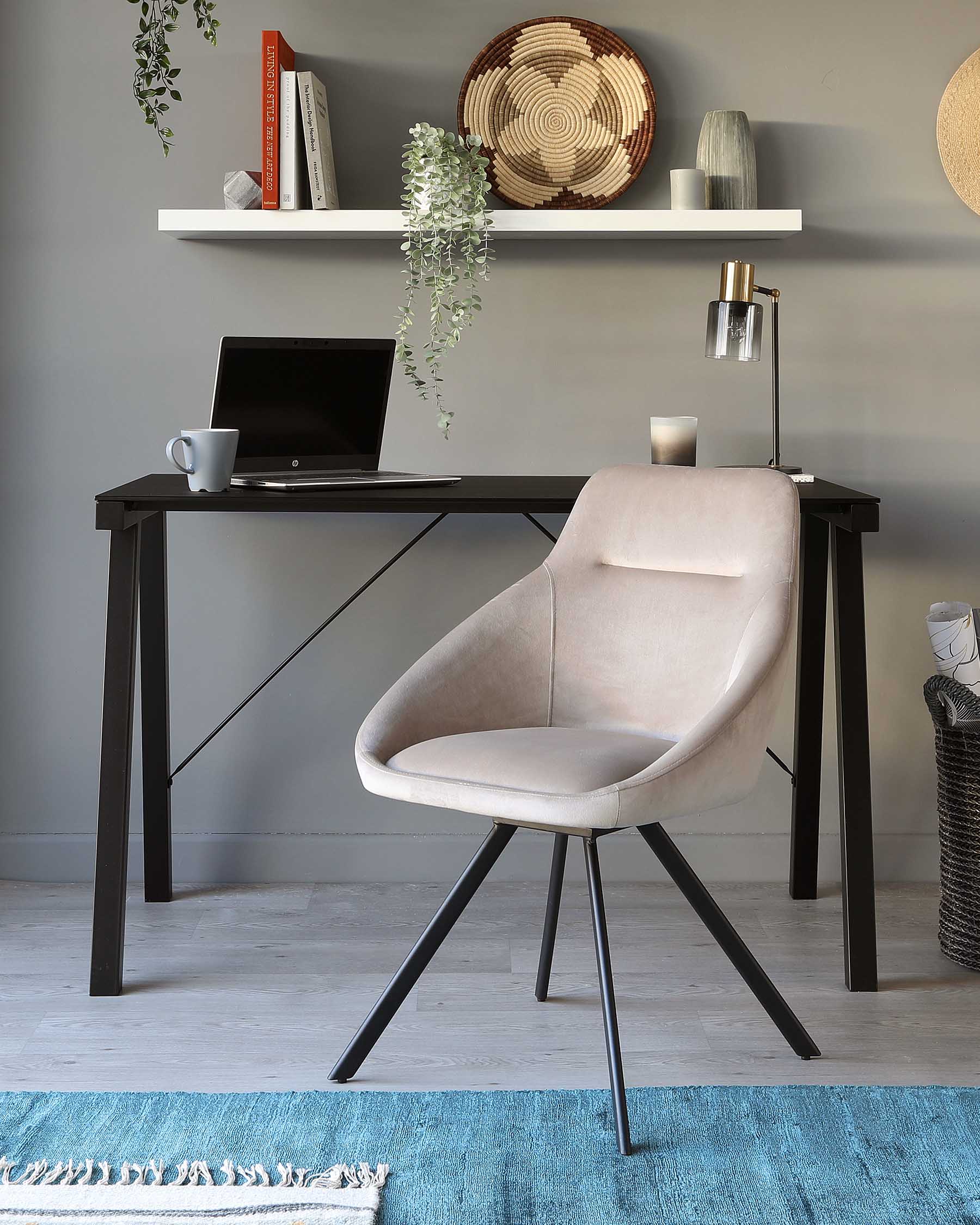 Modern minimalist home office setup featuring a sleek black writing desk with a straight silhouette and diagonal bracing, paired with a contemporary light beige upholstered chair with a curved back and tapered black metal legs. The setting is enhanced by a floating white shelf with decor above the desk.