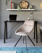 Contemporary furniture set against a grey wall, featuring a sleek black writing desk with a crossbeam support and a modern beige upholstered chair with black metal legs. The scene is accented by a floating white shelf above, adorned with decorative items and a hanging green plant. A stylish table lamp, decorative wicker plate, and books also complement the workspace setup.