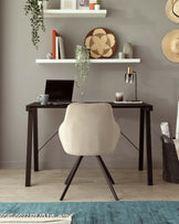 Modern home office setup featuring a sleek, dark wood desk with a minimalist design, paired with a beige upholstered chair with a curved backrest and angled metal legs. A floating white shelf is mounted above the desk, accessorized with decorative items and books.