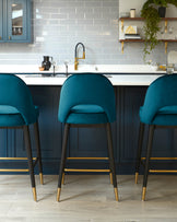 Three modern bar stools with plush teal upholstery and dark wooden legs accented with golden tips and crossbars.