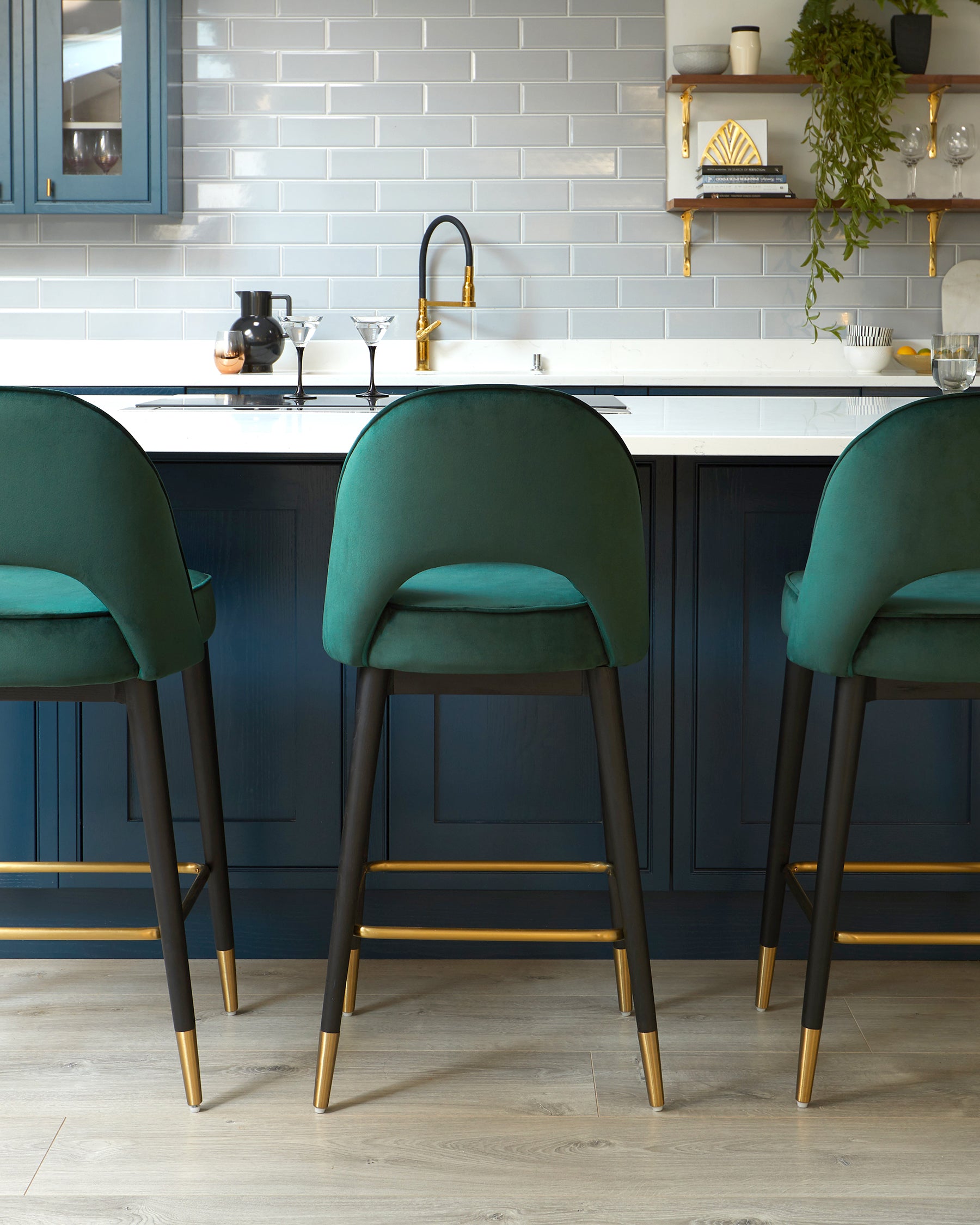 Three elegant teal upholstered bar stools with slim dark wooden legs, featuring brass accents at the tips and footrests, positioned in front of a kitchen counter with a dark blue base.