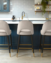 Three modern bar stools with elegant dark wood frames and gold-coloured footrests, featuring plush, light grey upholstery and curved backrests.