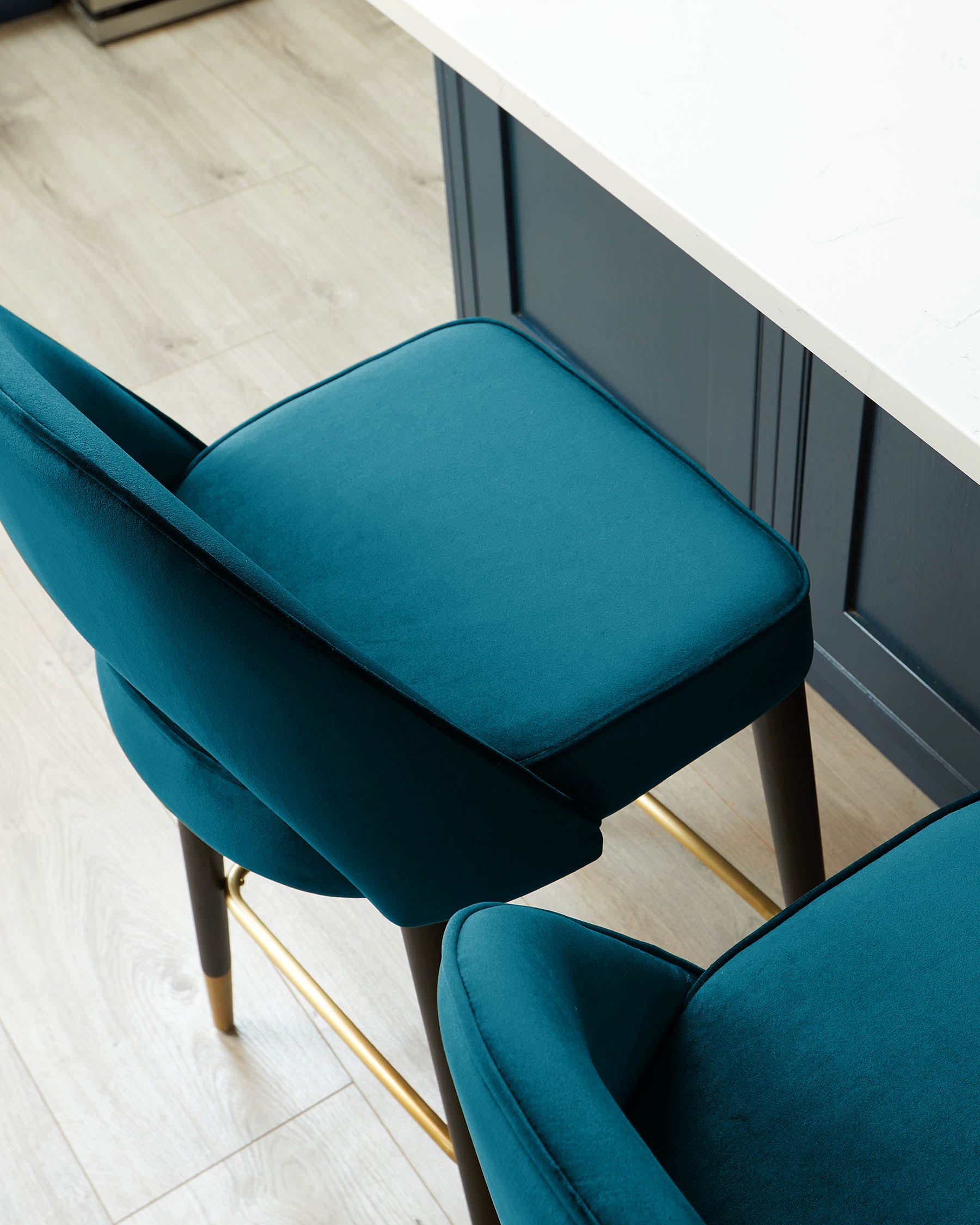 Elegant teal velvet dining chair with sleek brass legs and comfortable cushioned seating, set next to a modern white tabletop and against a grey panelled wall on a light wooden floor.