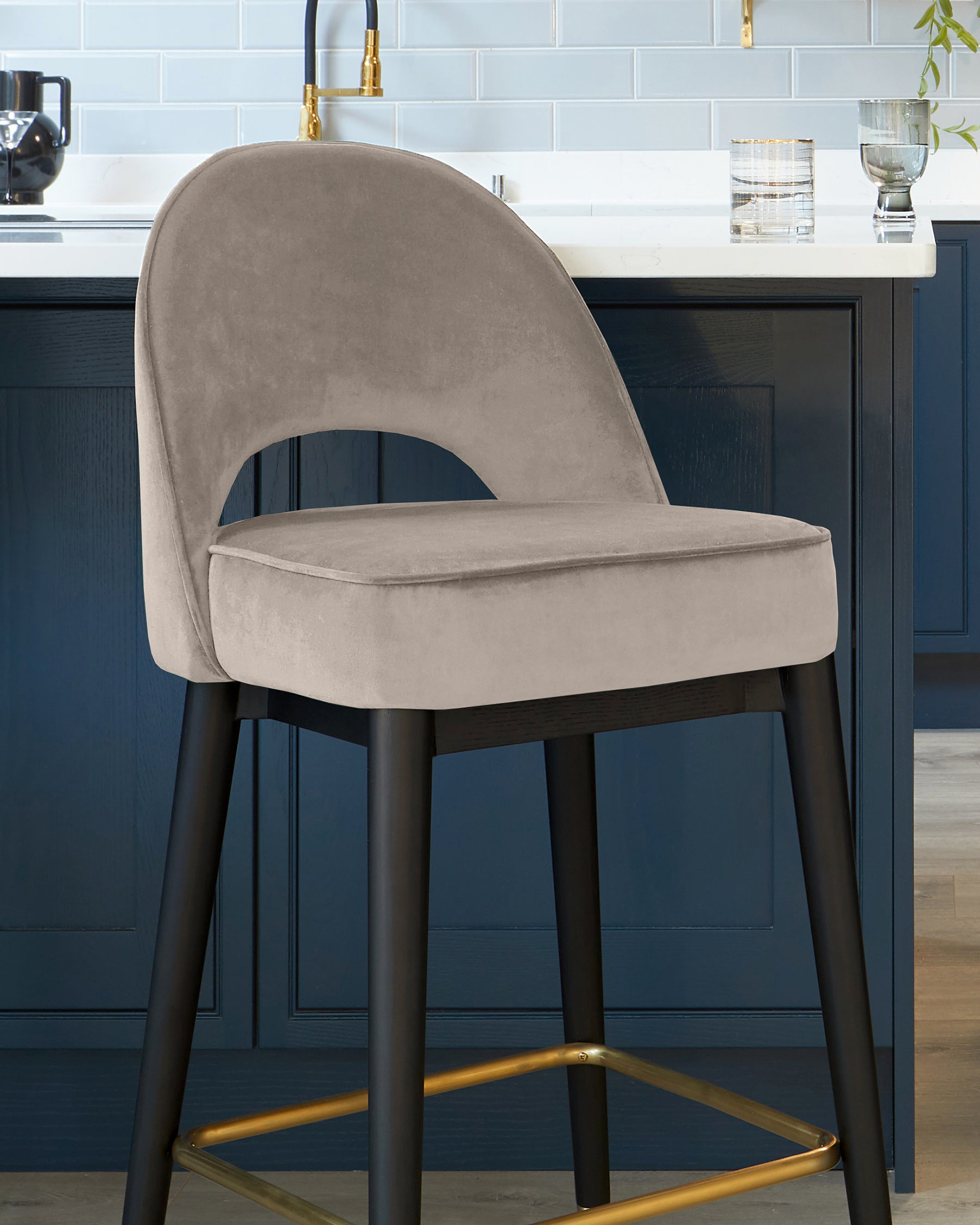 A modern velvet upholstered bar stool with a curved backrest and a cut-out detail, featuring sleek black legs with gold footrest accents, positioned against a kitchen island with blue cabinetry.