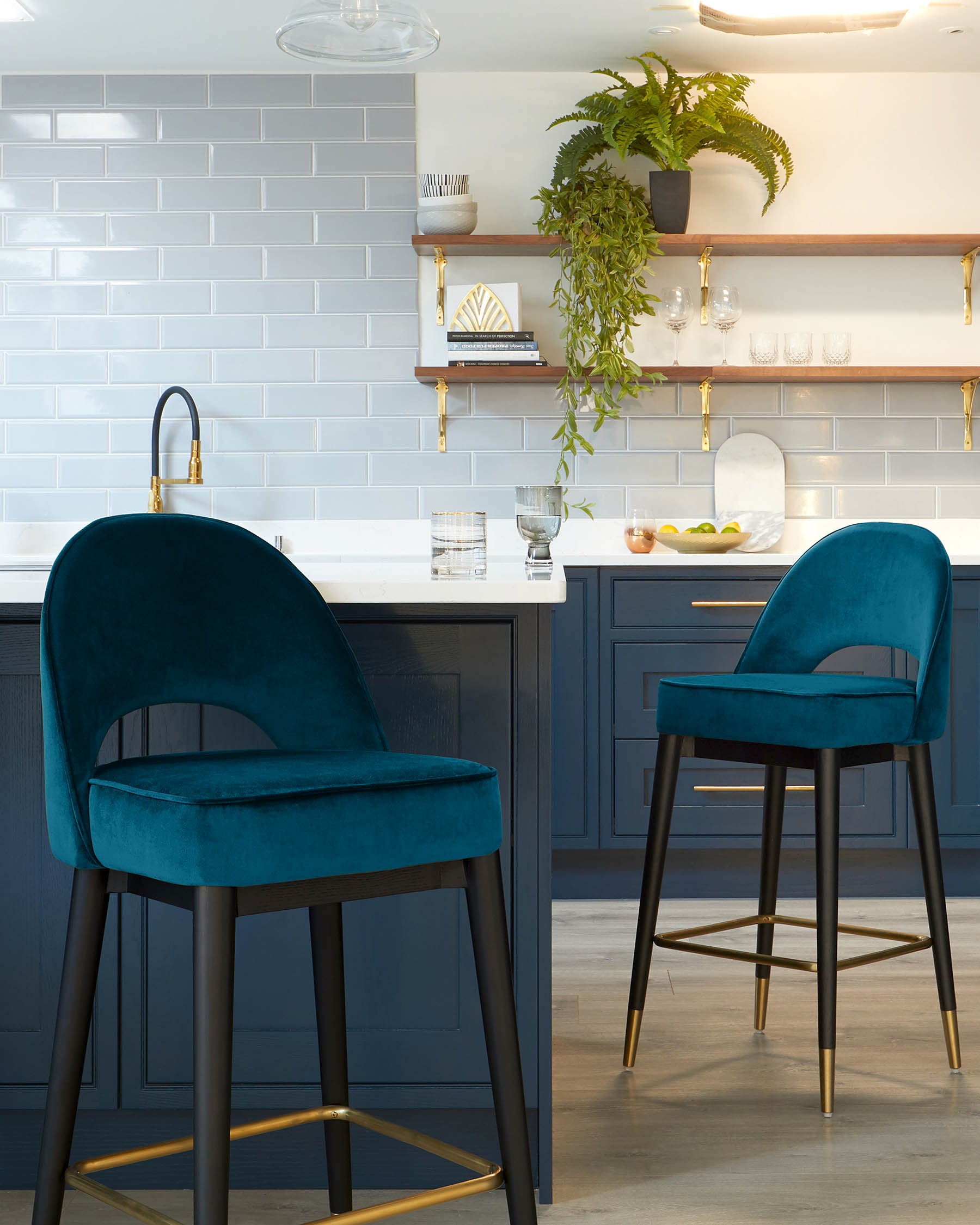 Two elegant bar stools with plush teal velvet upholstery and curved backrests. Both stools feature sleek black legs with golden footrest accents, adding a touch of luxury to the modern design.