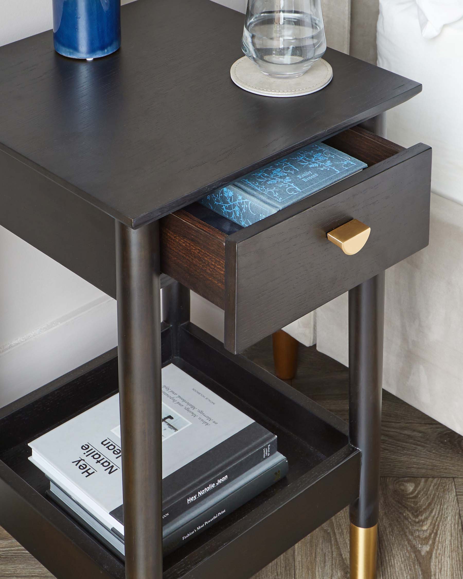 A contemporary dark wood nightstand featuring a smooth finish, a single drawer with a brass handle, and an open lower shelf. The legs are slender and partially accented with a golden tone at the bottom. On the nightstand, there is a decorative blue vase, a clear glass of water resting on a coaster, and a selection of books stored on the shelf below.