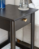 A modern dark-stained wooden nightstand with a rectangular top, featuring clean lines, a single drawer with a golden knob, and four straight, square legs creating an open base. There are items resting on the tabletop, including a carafe of water on a coaster and a decorative blue vase. The nightstand is positioned next to a light-coloured upholstered bed.