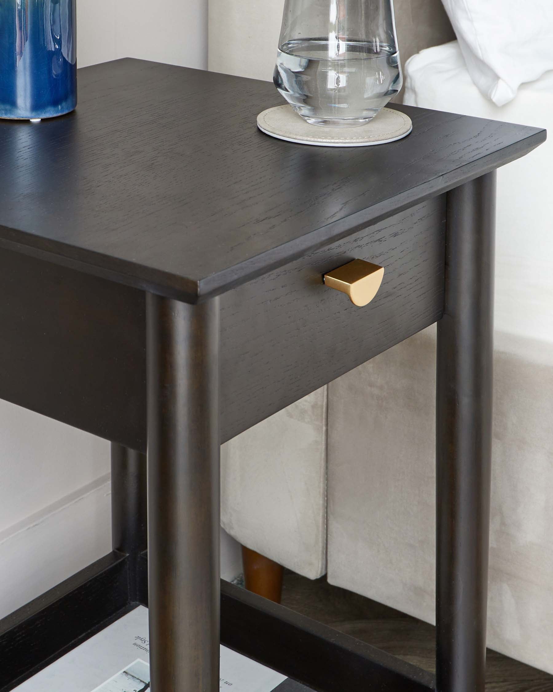 A modern dark-stained wooden nightstand with a rectangular top, featuring clean lines, a single drawer with a golden knob, and four straight, square legs creating an open base. There are items resting on the tabletop, including a carafe of water on a coaster and a decorative blue vase. The nightstand is positioned next to a light-coloured upholstered bed.