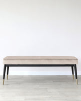 Modern beige upholstered bench with black wooden frame and brass tipped legs, set against a white wall and light wooden flooring.