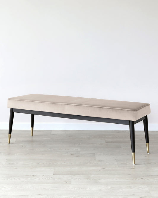 Elegant modern bench with a plush, light beige upholstered cushion on a dark wooden base with black and gold tapered legs, positioned on a light wooden floor against a white wall.