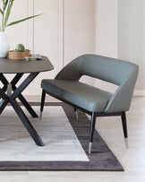 Modern minimalist style furniture set featuring a sleek, dark grey armchair with a curved backrest and metal-tipped legs, accompanied by an angular black dining table with a distinctive X-shaped base and metallic accents at the feet. Set on a two-tone grey area rug, the scene is completed with a decorative vase and wooden tray on the table.