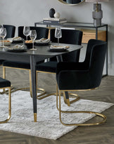 Modern dining room furniture set with a dark wood table featuring gold metal accents on the legs, surrounded by plush black velvet chairs with curved backrests and gold metal framework on the base. A sleek black and gold console table is visible in the background, accessorized with decorative items. The ensemble is placed on a soft, textured white area rug.