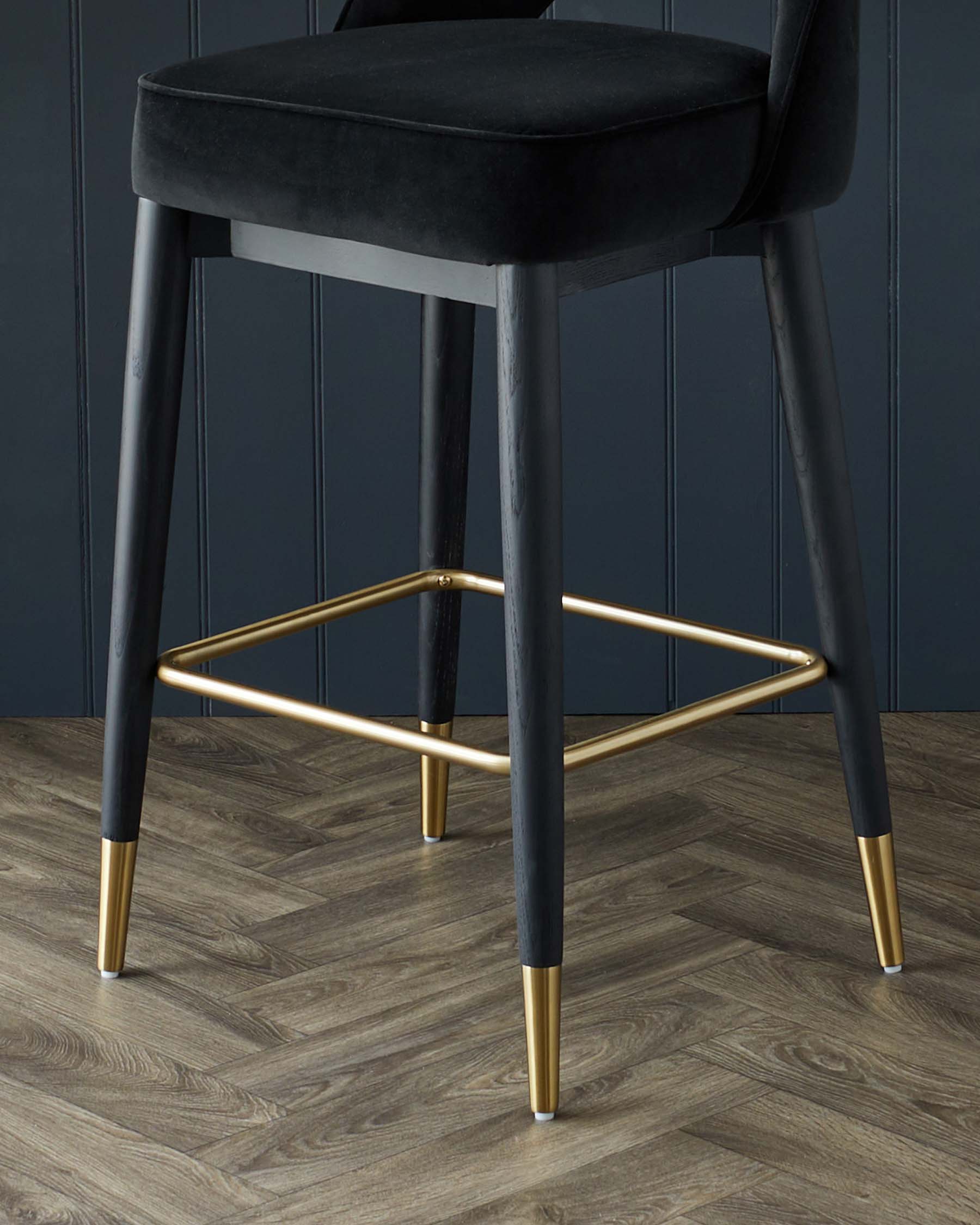 Elegant modern bar stool with black velvet cushioning and dark wooden legs accented with gold-coloured metal tips and a matching circular footrest. The stool is set against a dark blue wall and on a herringbone-patterned wooden floor.