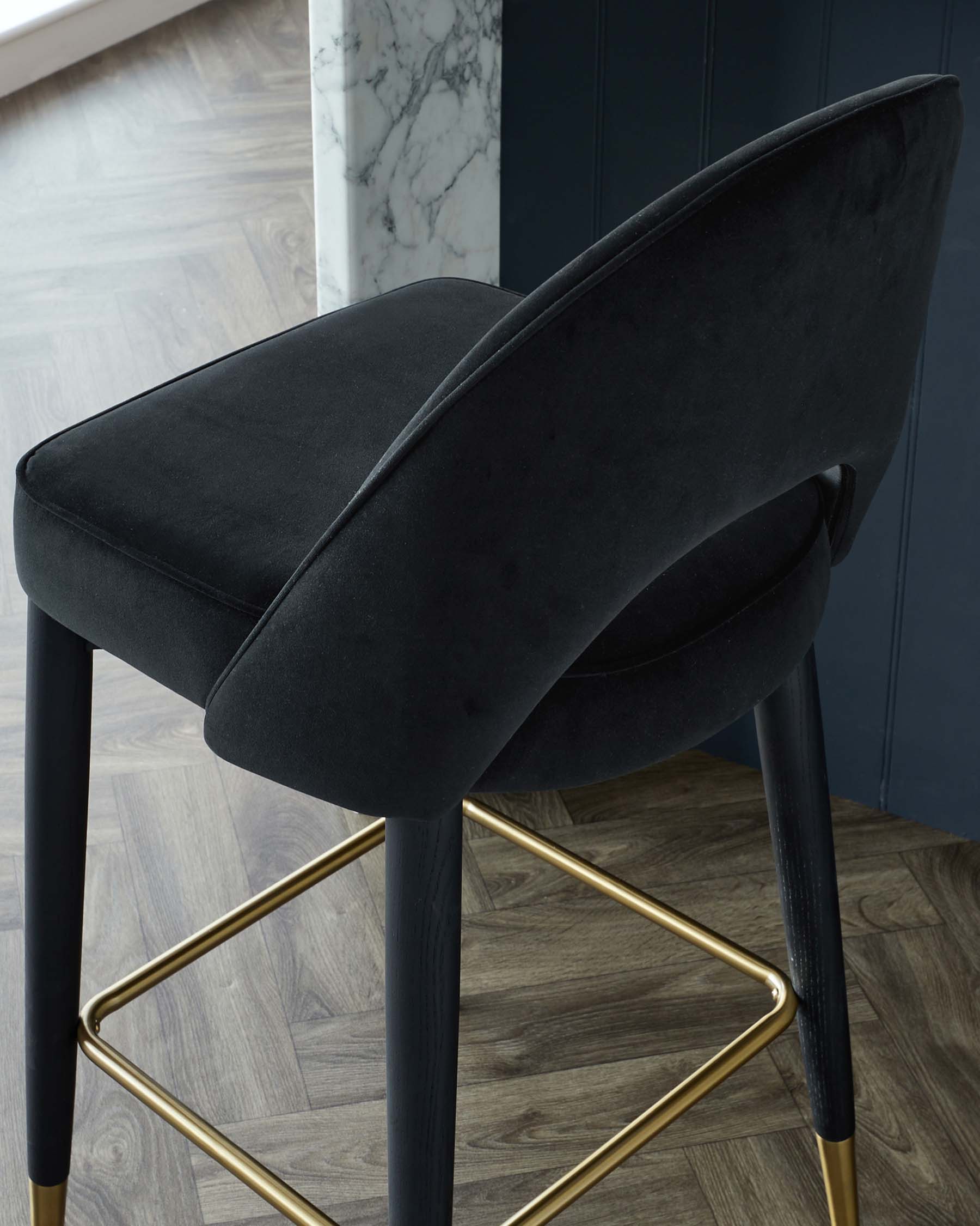 Elegant modern bar stool with black velvet upholstery, featuring a curved open backrest, a comfortable padded seat, and a sturdy frame with black and gold brass-finished metal legs, set against a backdrop of wood flooring and a marble accent wall.