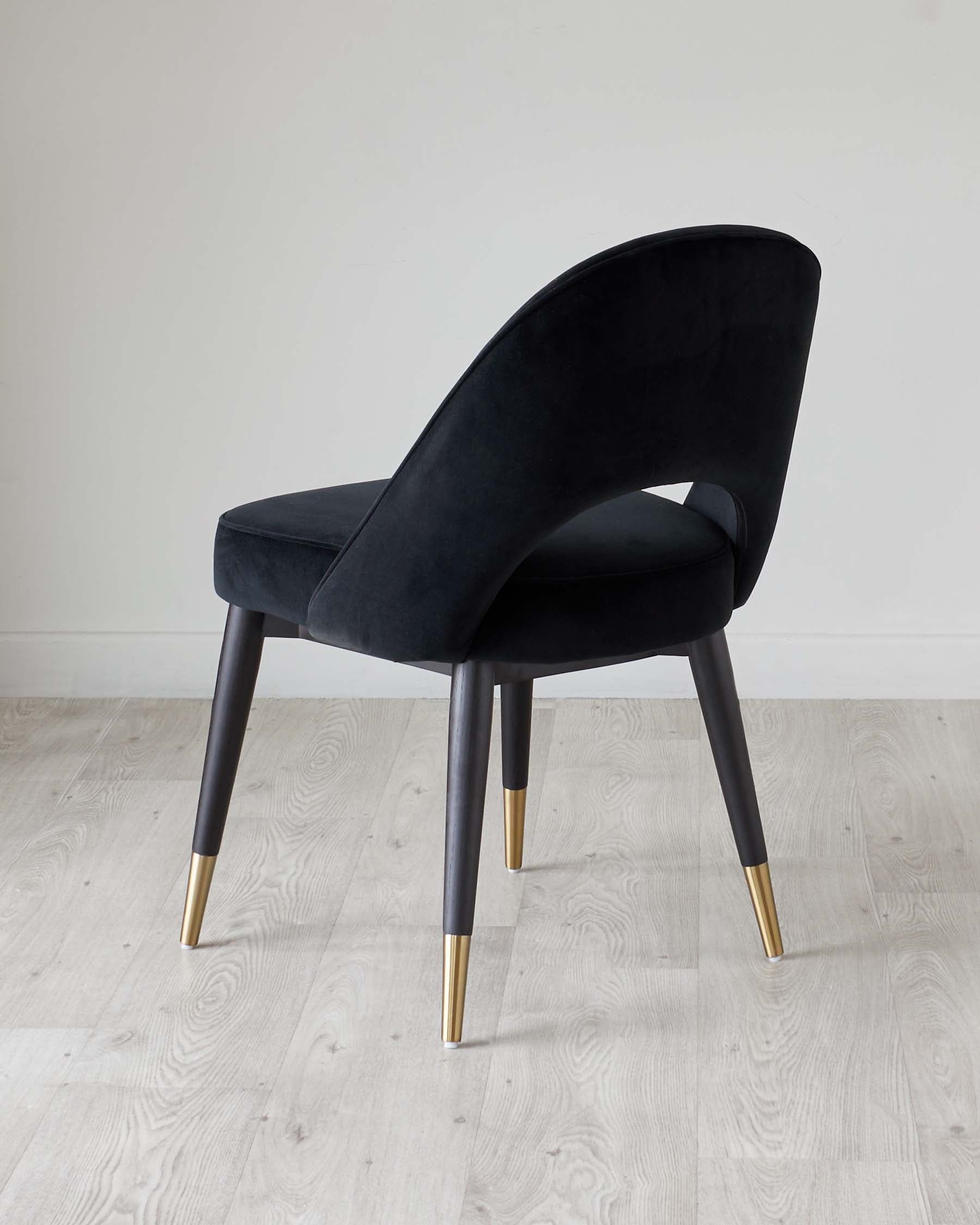 Elegant modern chair with a curved backrest and plush black velvet upholstery, featuring contrasting dark legs with chic gold tips, displayed on a light wooden floor against a neutral white wall backdrop.