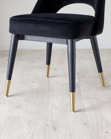 Elegant modern chair with black velvet upholstery and sleek black legs, accented with stylish golden tips, set against a light wooden floor.