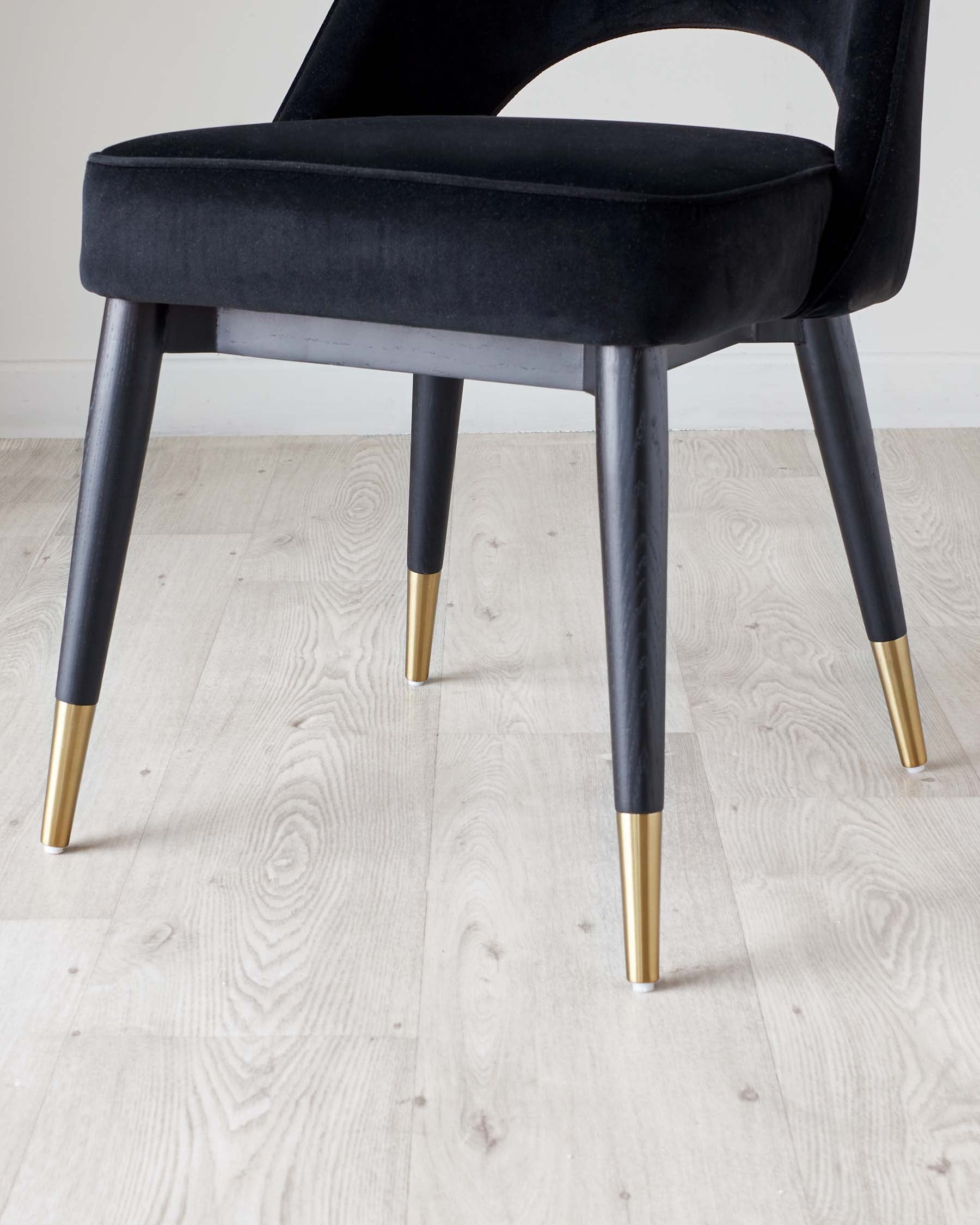 Elegant modern chair with black velvet upholstery and sleek black legs, accented with stylish golden tips, set against a light wooden floor.