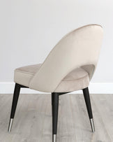 Elegant contemporary chair featuring a curved backrest and a cushioned seat upholstered in a light beige fabric, with a unique design that tapers down smoothly to merge with the back legs. The chair has contrasting black wooden legs with metallic silver tips, set against a light wooden floor and a plain white background.