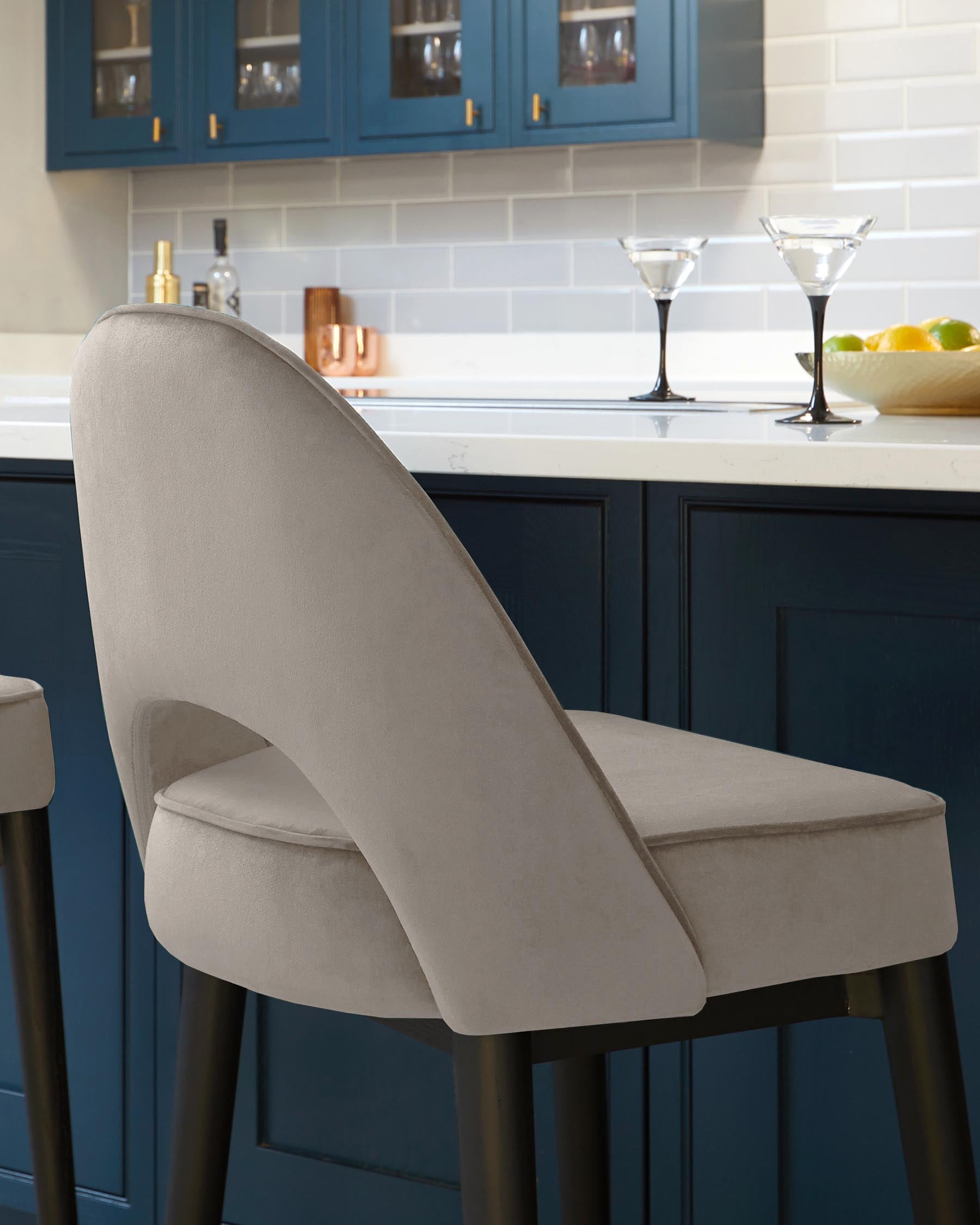 Elegant modern barstool with a curved backrest and plush seat cushion, upholstered in light beige fabric, juxtaposed against a kitchen island with dark blue cabinetry and white countertop.