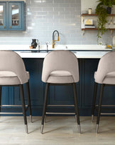 Elegant modern kitchen with three plush, upholstered bar stools featuring smooth curved backs and slender black legs with metallic tips, positioned at a white countertop.