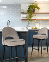 Two modern upholstered bar stools with a sleek, curved backrest design, a comfortable padded seat in a light grey fabric, and a sturdy black metal frame with four legs and a circular footrest.