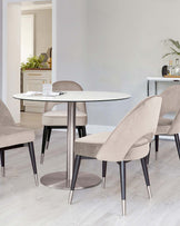 Modern round dining table with a white marble-like tabletop and a single central metal support on a circular base, surrounded by four contemporary upholstered chairs in a soft beige fabric with black tapered wooden legs accented with metal tips. The setting is a bright, minimalist room with wood flooring.