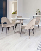 A modern dining set featuring a round white tabletop with a central metallic base and four surrounding chairs upholstered in a taupe velvet fabric with black and metallic-tipped legs on a light hardwood floor.