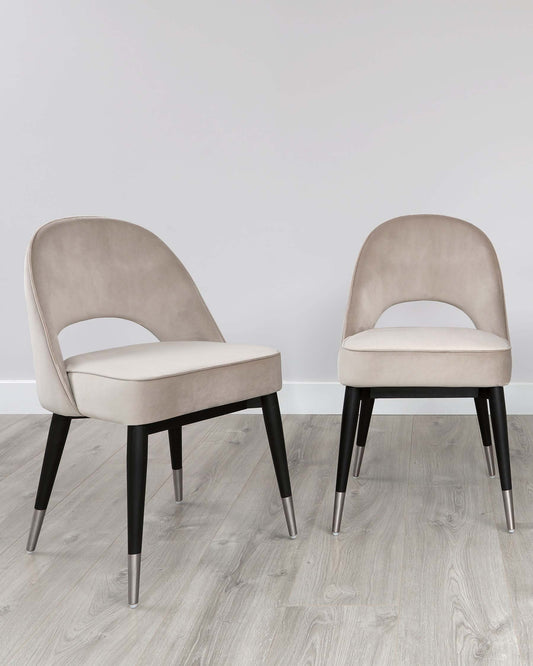 Two contemporary-style dining chairs with beige velvet upholstery and contrasting black legs with metallic tips, positioned on a grey wood-effect floor against a neutral wall.