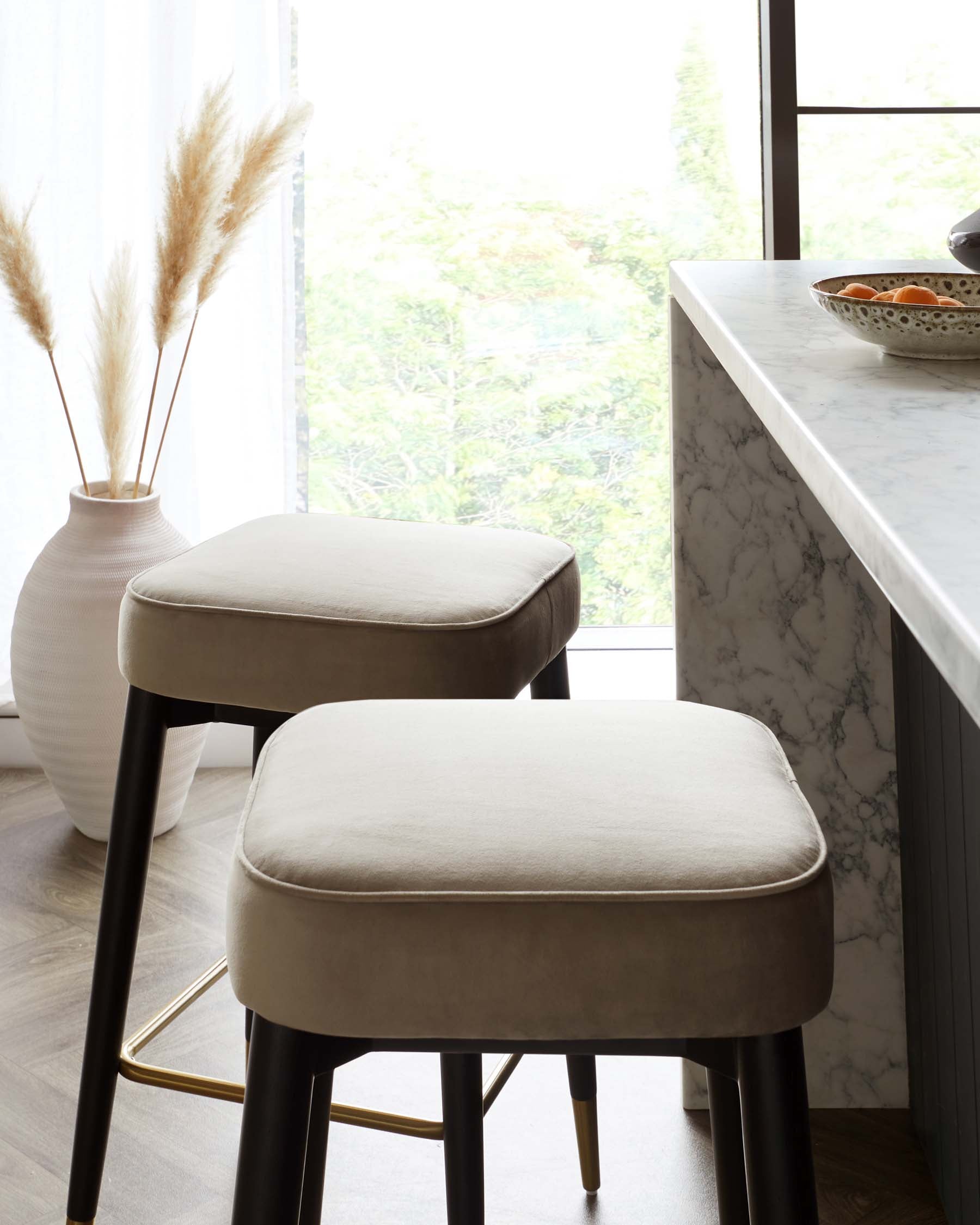 Two modern minimalist bar stools with soft, grey upholstery on the seat cushions, and sleek black metal legs that include a hint of gold at the bottom, positioned next to a white marble countertop.