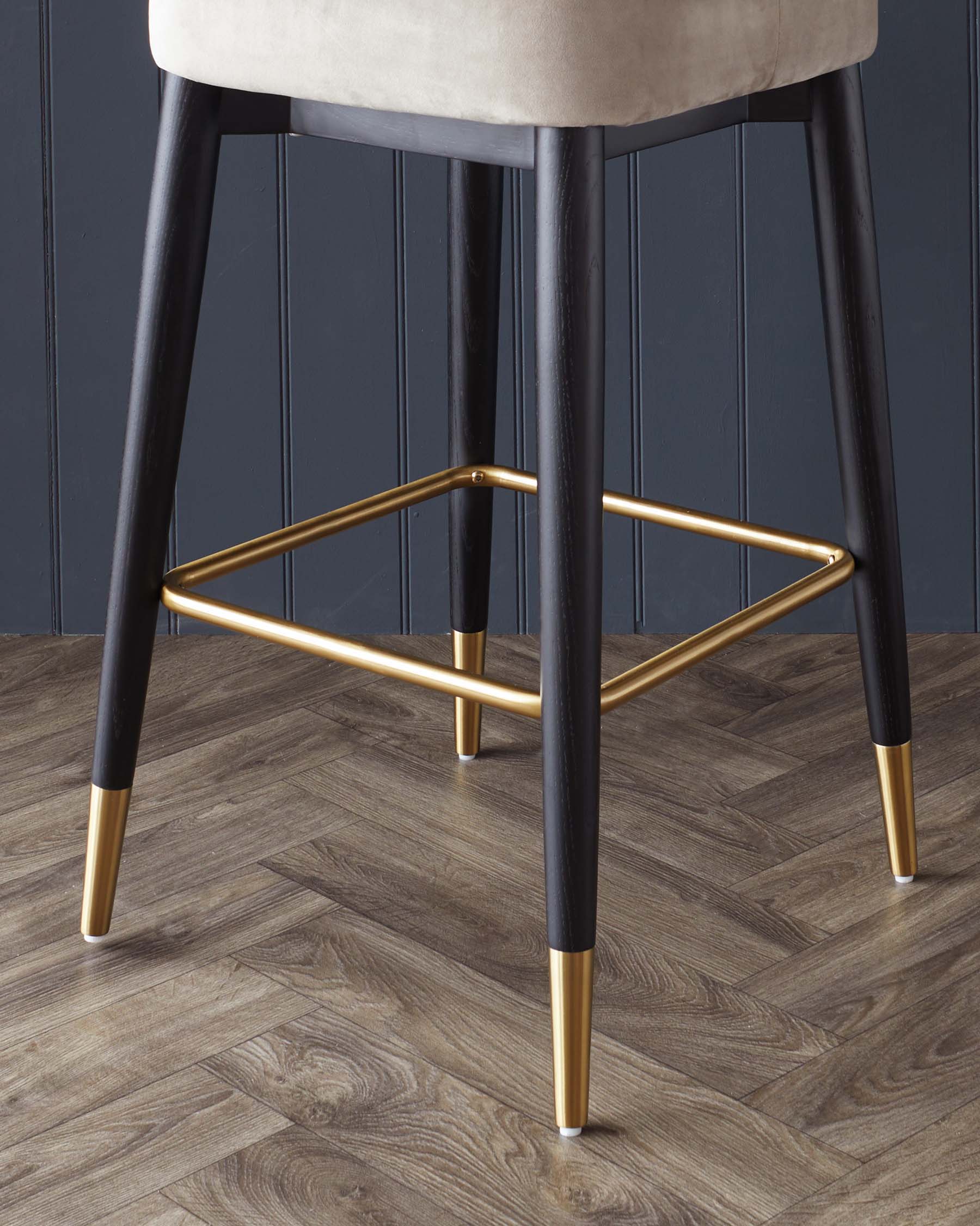 Modern bar stool featuring dark tapered legs with gold-finished tips and footrest, complemented by a plush, light upholstered seat against a dark panelled wall and herringbone wood flooring.