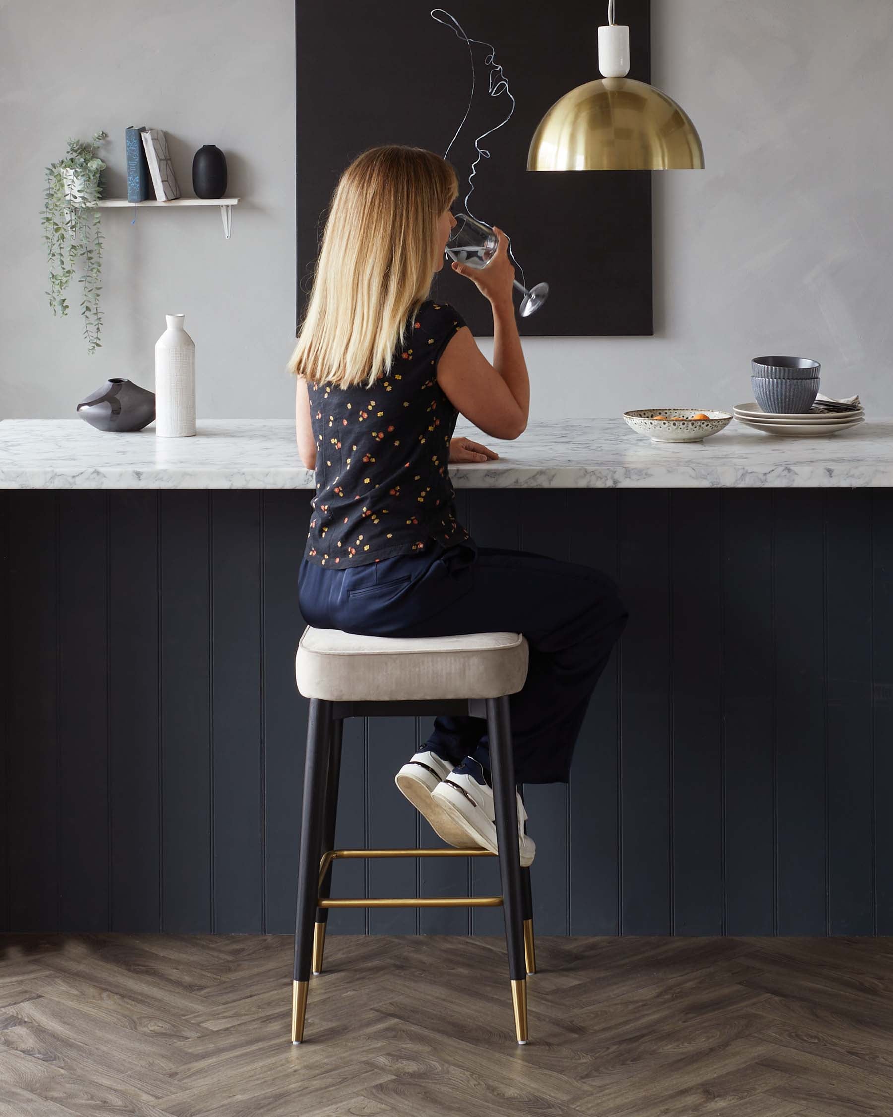 Modern counter-height bar stool with dark wooden legs, gold footrest, and a cushioned beige seat. A sleek marble countertop can also be seen.