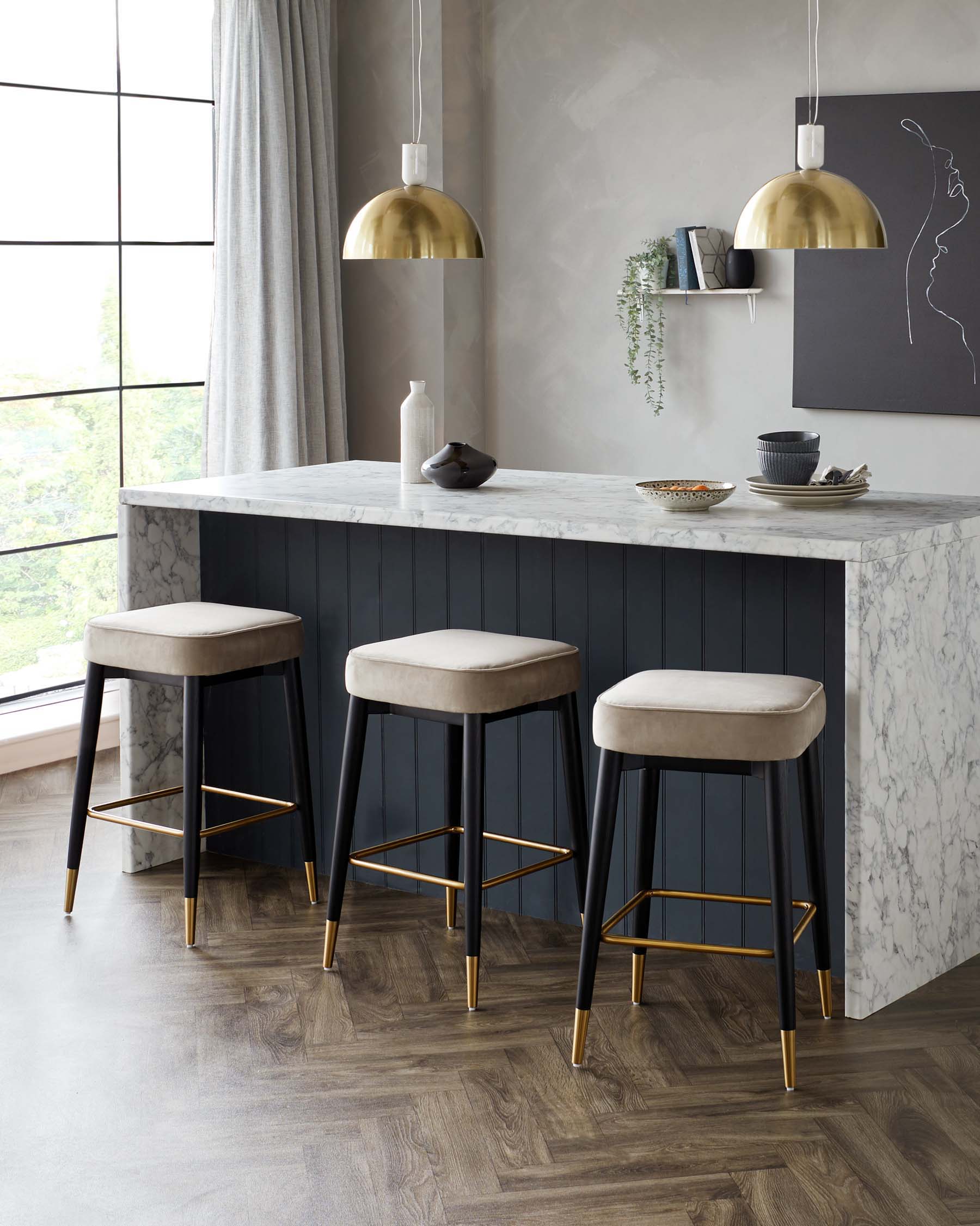 Three modern bar stools with black frames and brass accents, featuring round light-coloured upholstered seats, situated against a kitchen island with a marble countertop.