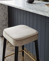 Modern bar stool with a beige velvet seat cushion, black legs, and metallic gold footrest details, in front of a kitchen island with a marble countertop and black wooden base.