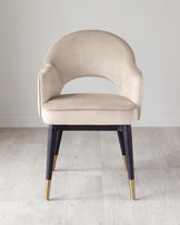 Elegant modern accent chair with a curved back, upholstered in a light beige velvet fabric, and supported by four tapered legs in a deep blue with golden tips, set against a light hardwood floor and a pale grey background.