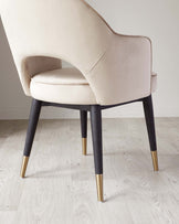 Elegant modern chair featuring a cream upholstered seat and backrest with a smooth curved design and visible seam detailing. The chair stands on four angled solid legs in a dark finish, accented with gold-toned metal tips at the bottom, set against a light wood floor.