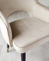 Elegant modern chair with a curvaceous beige velvet upholstered seat and back, featuring a smooth finish and subtle stitching detail, complemented by tapered dark wooden legs with brass-capped feet.