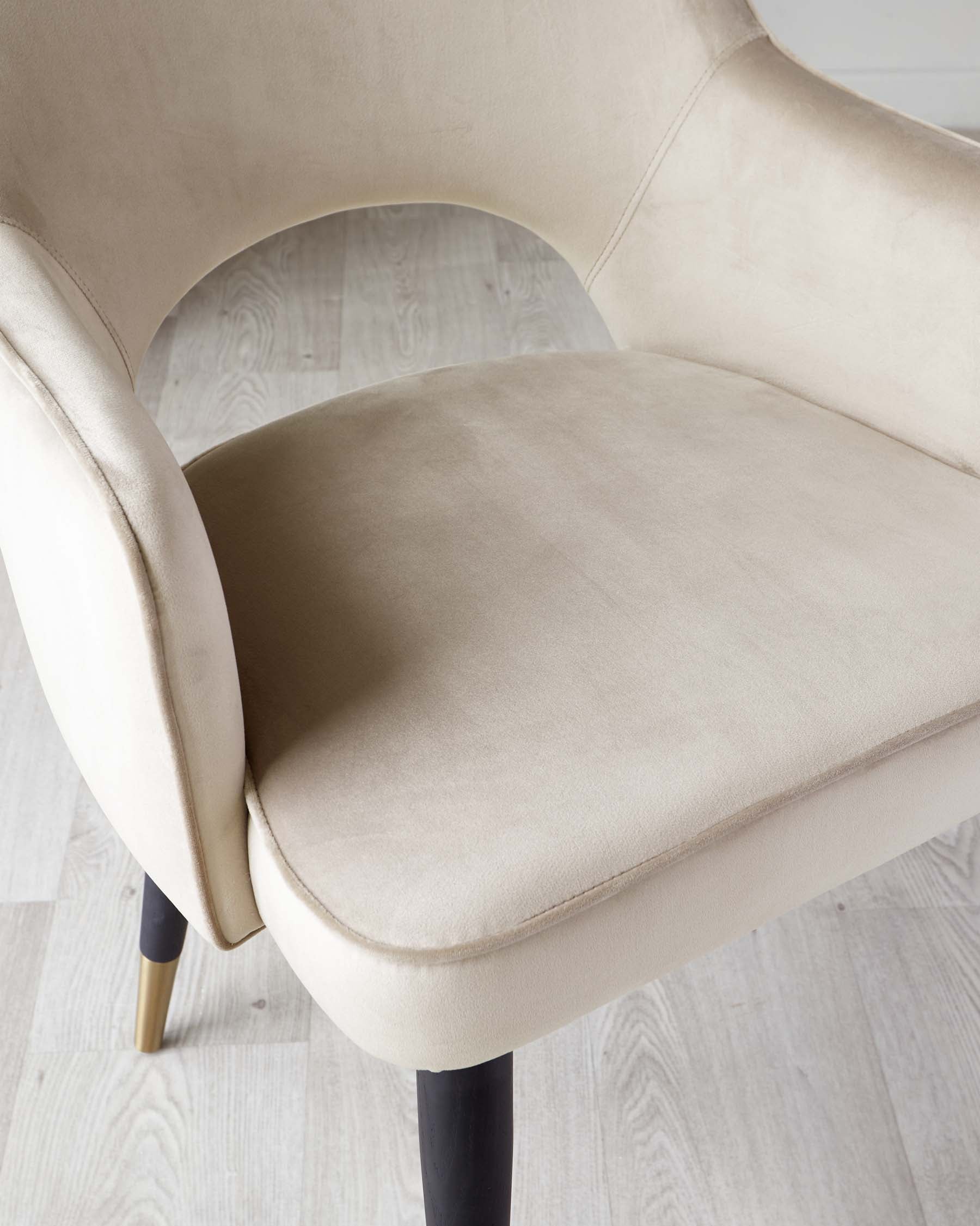 Elegant modern chair with a curvaceous beige velvet upholstered seat and back, featuring a smooth finish and subtle stitching detail, complemented by tapered dark wooden legs with brass-capped feet.