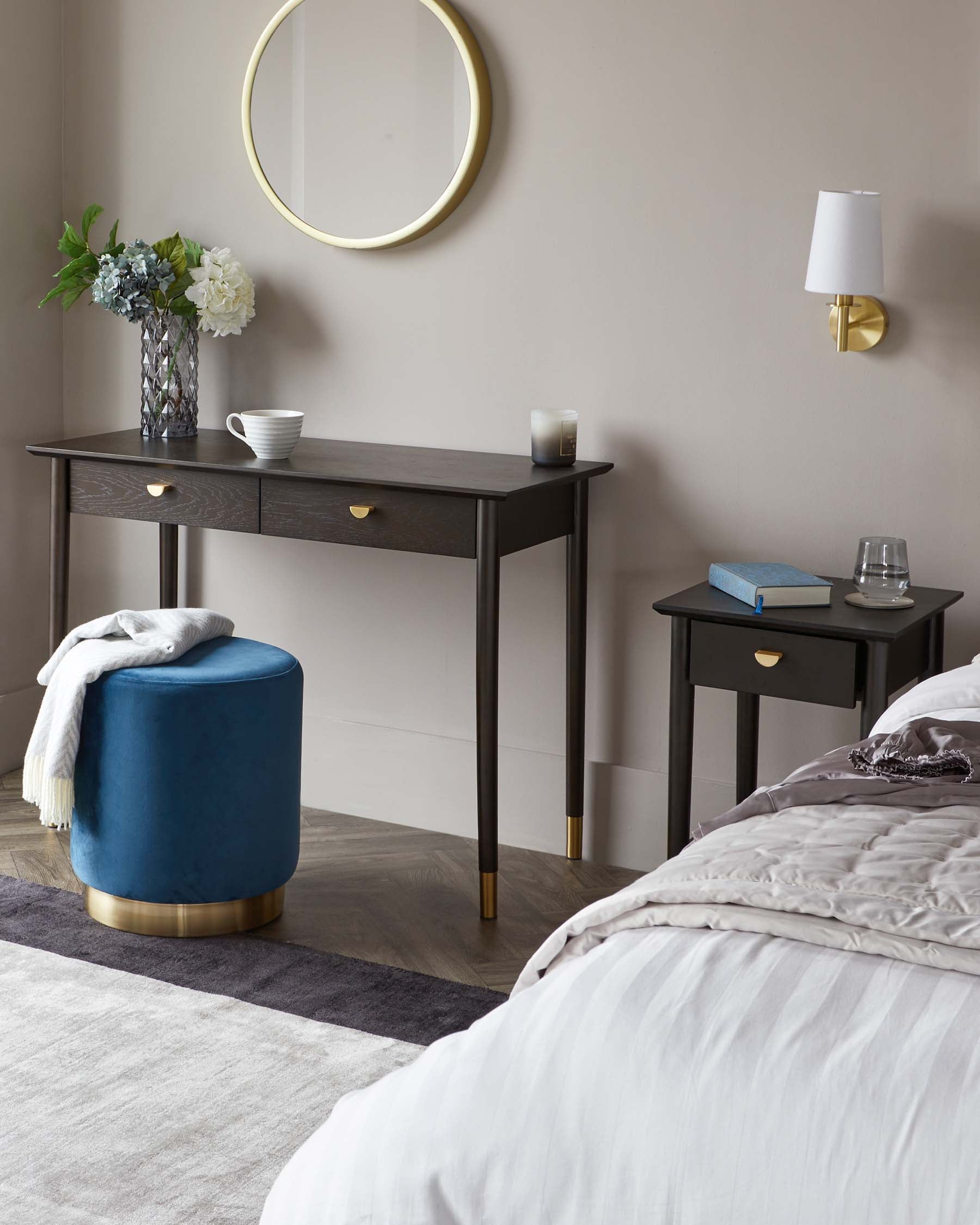 Elegant bedroom furniture set including a dark wooden console table with brass handles and slender legs, a matching dark wooden side table with a single drawer and brass accents, and a luxurious round velvet ottoman in deep blue with a brass base. A contemporary wall-mounted lamp and circular gold-framed mirror complement the ensemble.