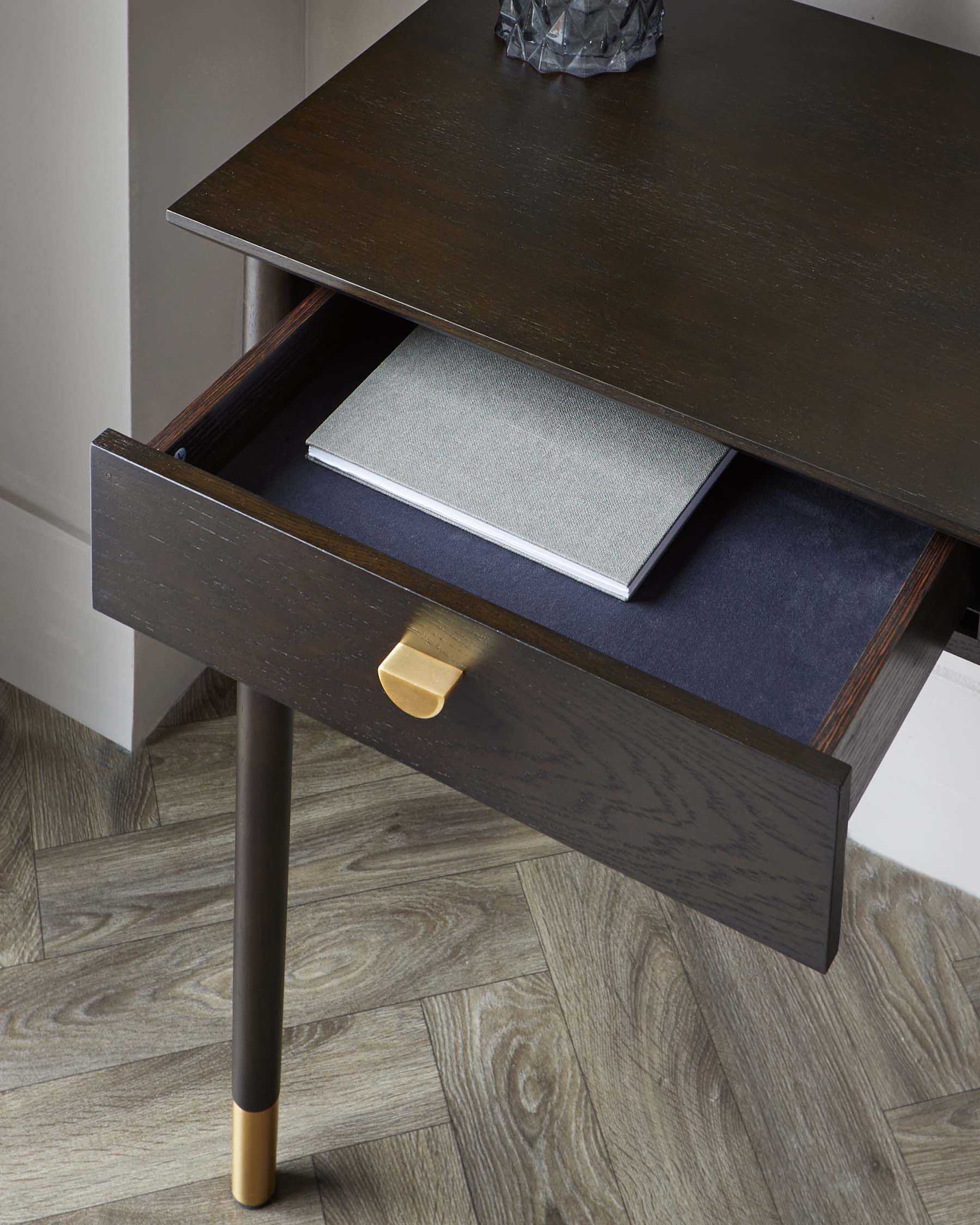 Modern dark wooden desk with an open drawer revealing books and an elegant brass handle, supported by slim legs tipped with brass accents on a herringbone wood floor.