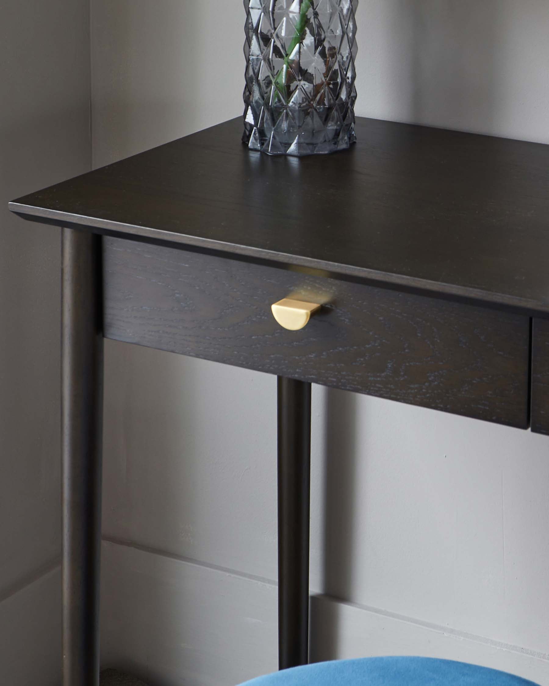 Contemporary dark brown wooden console table with a sleek top surface, featuring a single visible drawer with a brass-coloured, half-moon pull handle. The table stands on straight, round metal legs, providing a modern contrast to the wooden elements. A decorative glass vase resides atop the table.