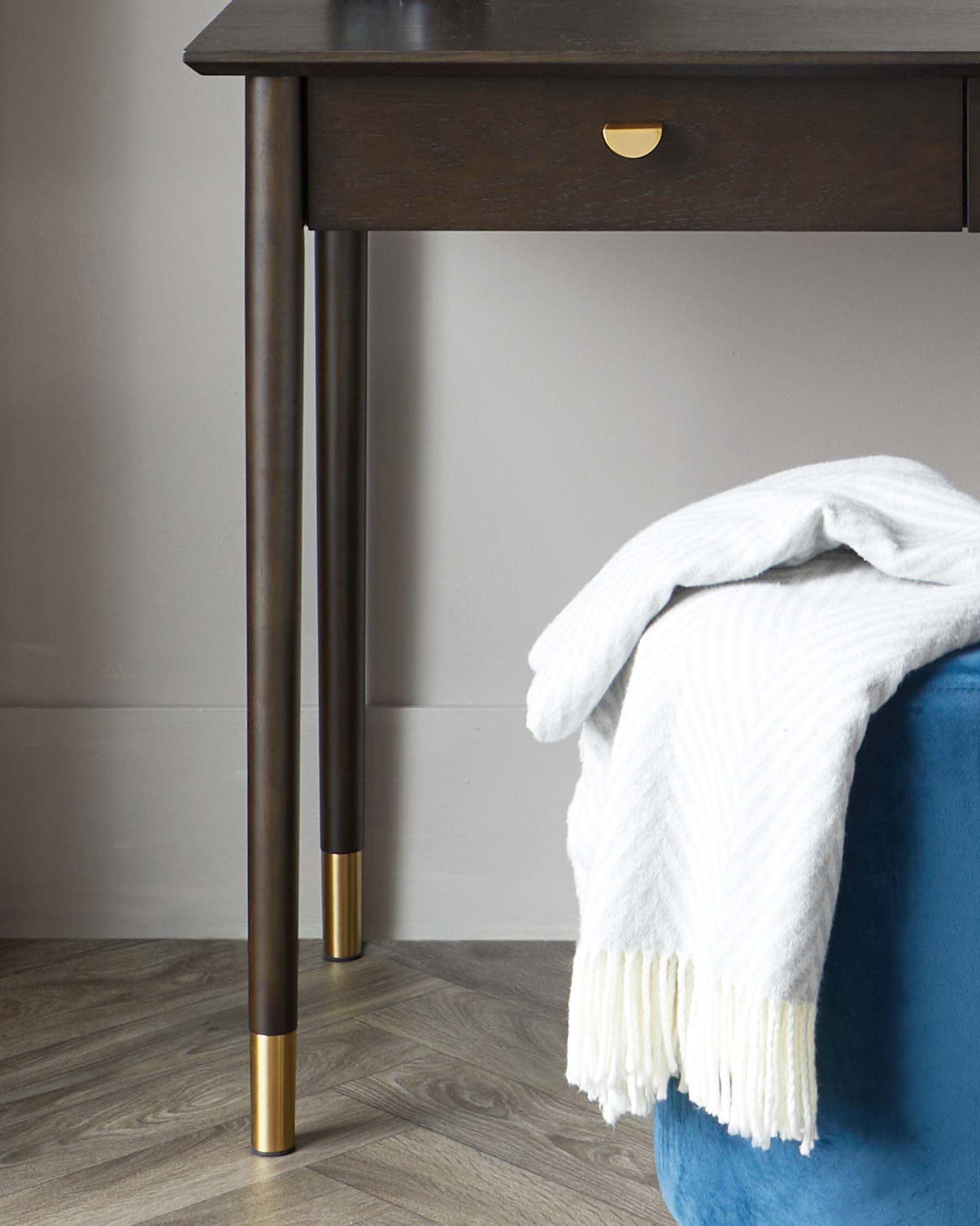 Contemporary espresso brown side table with a single gold-coloured drawer pull and slender legs, complemented by brass-tipped feet. Adjacent to the table is a plush, royal blue velvet ottoman draped with a white, fringed throw blanket.